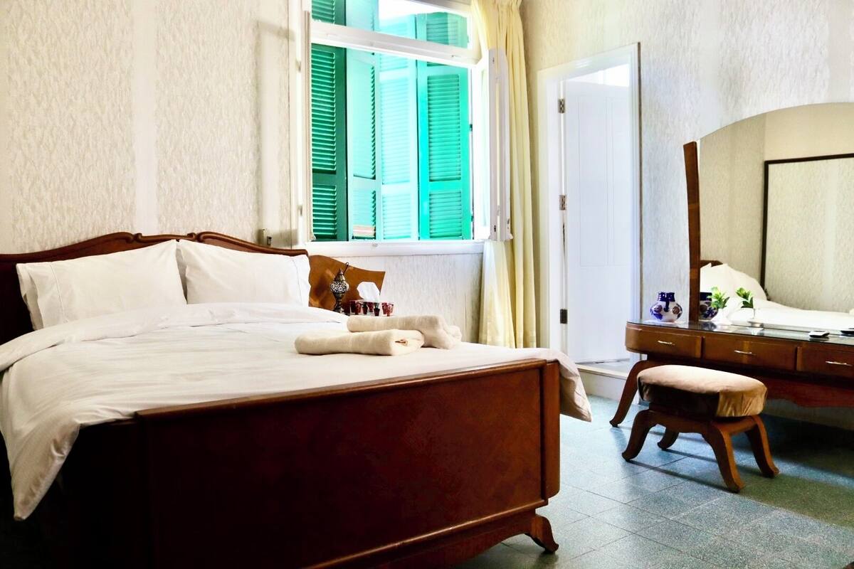 A hotel room with a wooden bed, a bedside table with a mirror, and a window with green shutters.