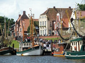 A harbor with several boats docked, a crowd of people on a dock, and buildings with red-tiled roofs in the background. - The "Kiebitznest" in Greetsiel meets high demands (Krummhoern)
