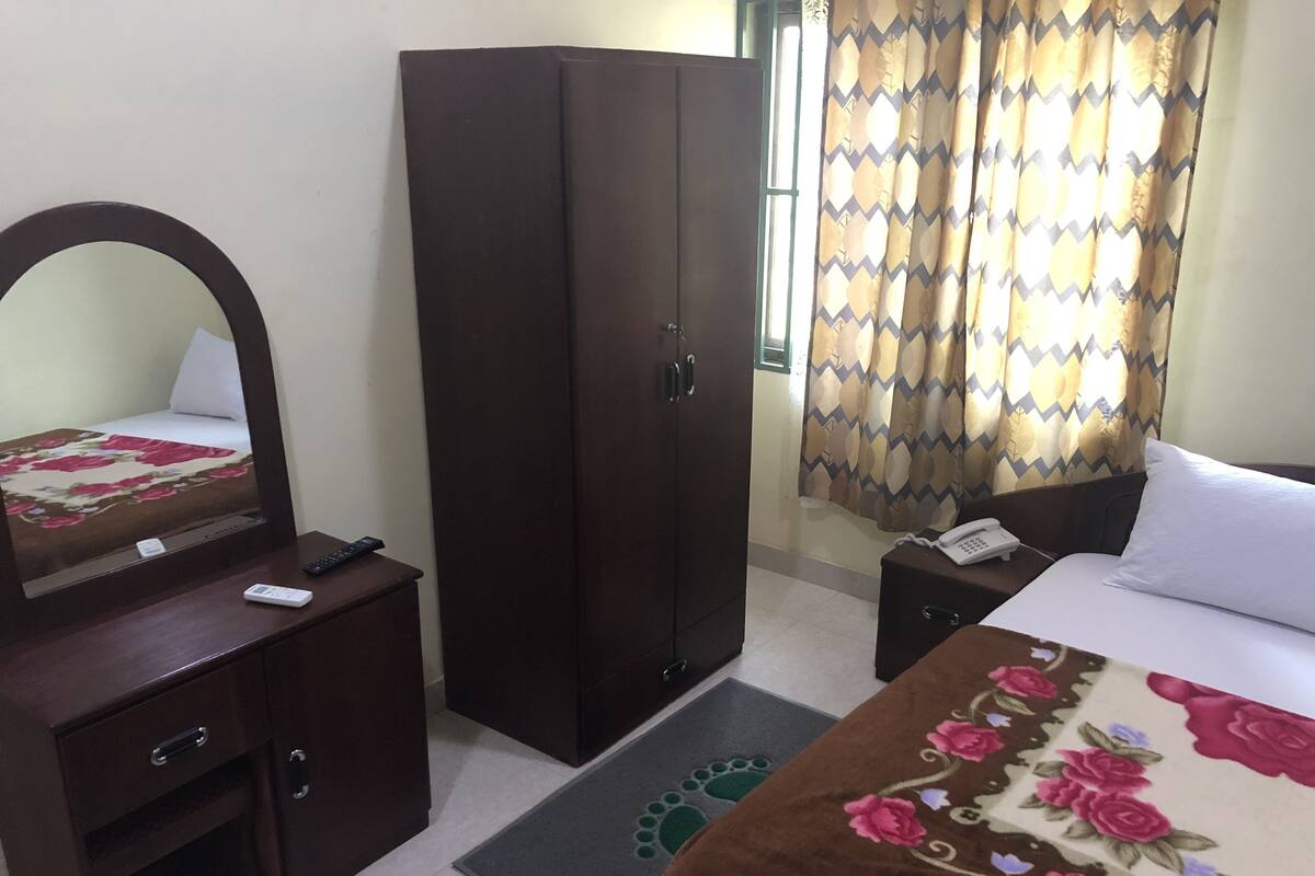 A hotel room with a bed, a wardrobe, a bedside table with a phone, a mirror, and a desk with a remote control.