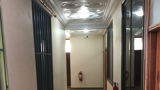 A hallway with a polished floor, a fire extinguisher mounted on the wall, and a window with metal bars on the left. - Ricardo's property (Accra)