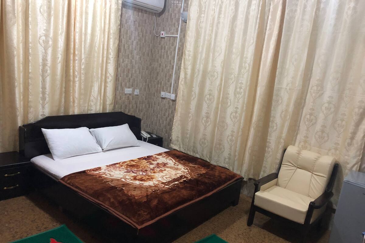 A hotel room with a bed, a chair, a nightstand, and a wall-mounted air conditioner.