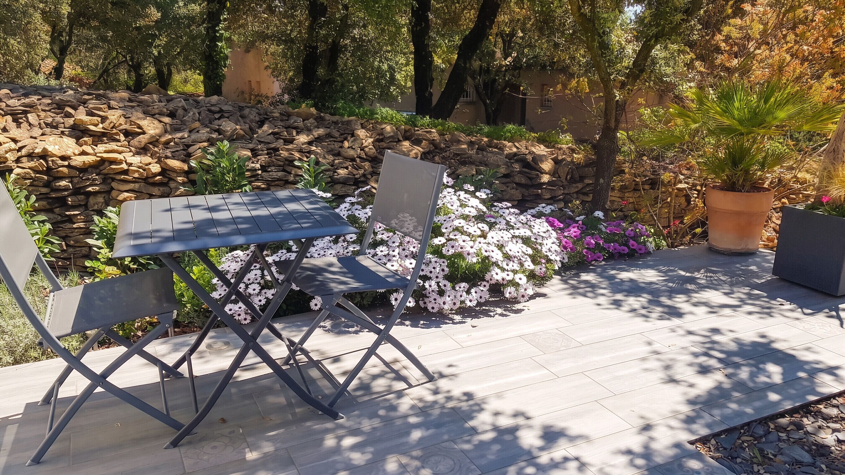 An outdoor seating area with a table and chairs, surrounded by a stone wall and various plants.