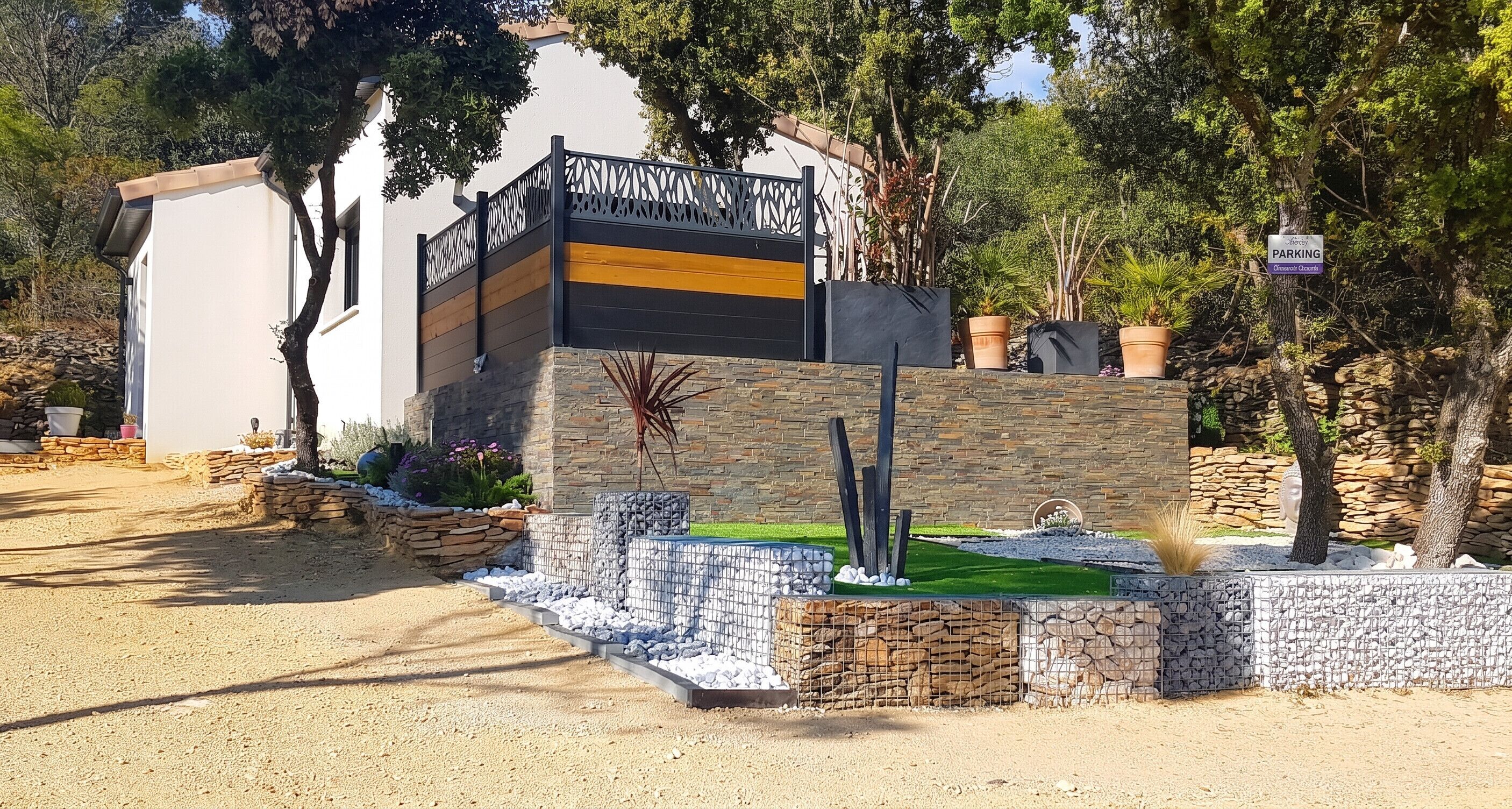 Un bâtiment moderne à la façade en pierre, un panneau indiquant un espace de stationnement et un jardin bien entretenu avec diverses plantes et un petit élément décoratif aquatique.