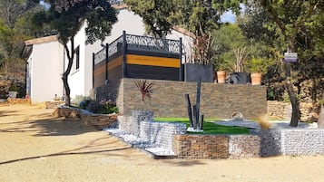 Un bâtiment moderne à la façade en pierre, un panneau indiquant un espace de stationnement et un jardin bien entretenu avec diverses plantes et un petit élément décoratif aquatique.