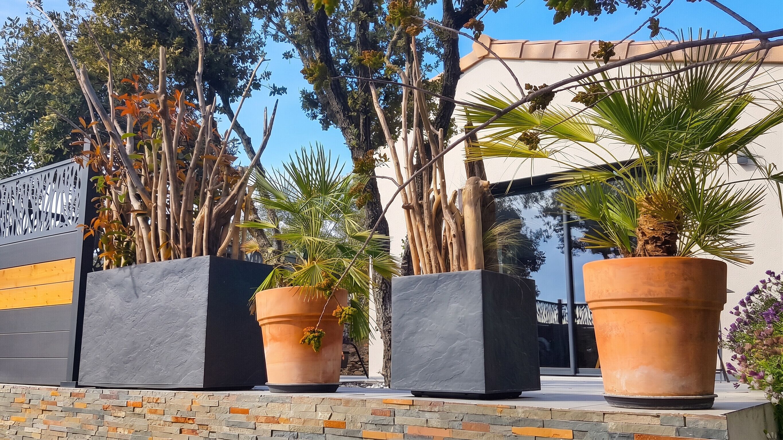 Une façade d’immeuble avec un mur en pierre, de grands pots de plantes et un portail en métal.