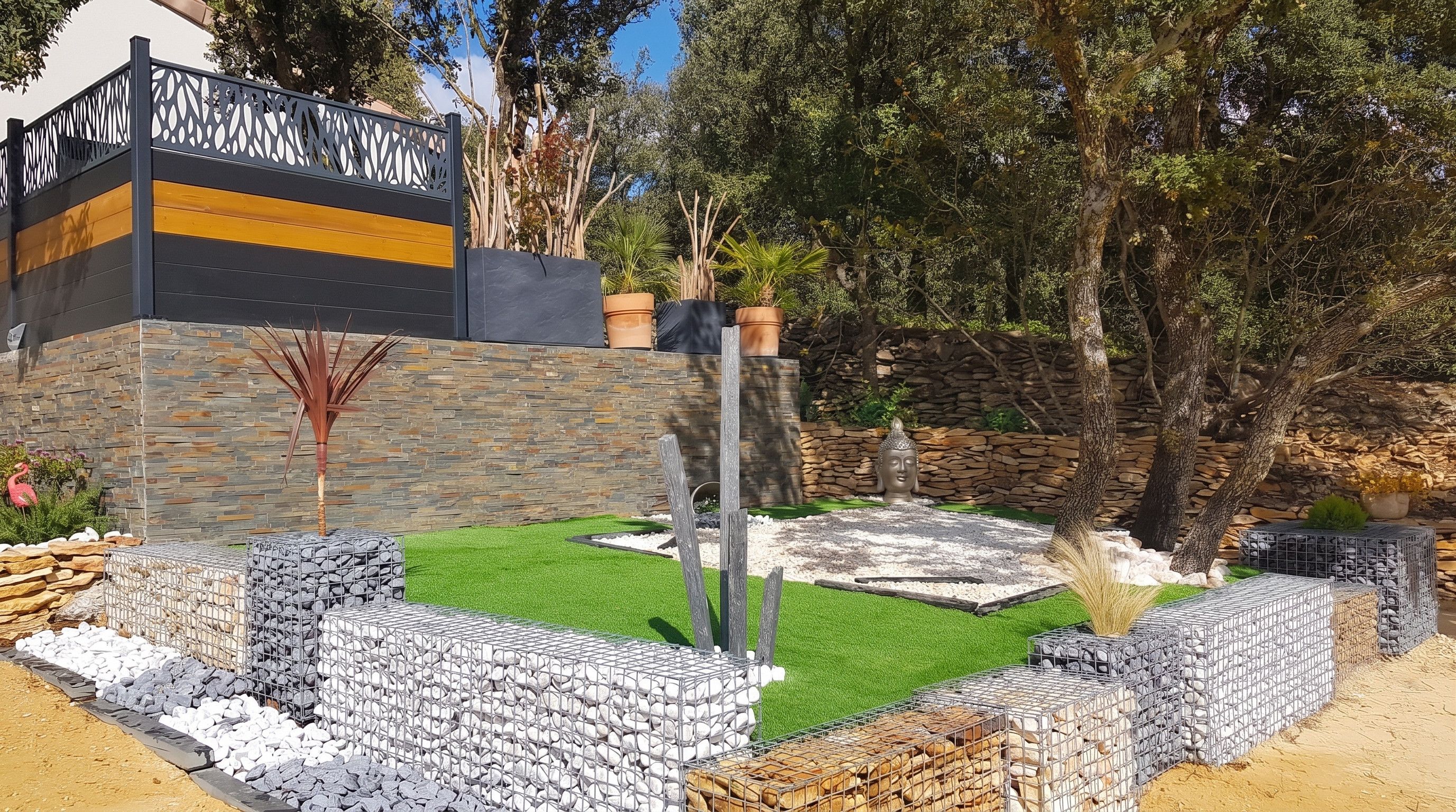 Un espace paysager aménagé en jardin, avec un mur en pierre, une statue de Bouddha et un panneau avec du texte.