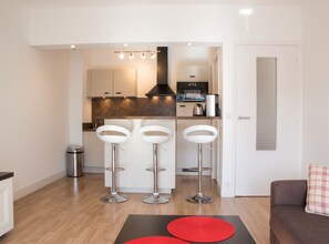 A modern kitchen with a built-in oven and a coffee maker. There are three white bar stools and a trash can. - Homerez - Apartment in Antibes (Antibes)