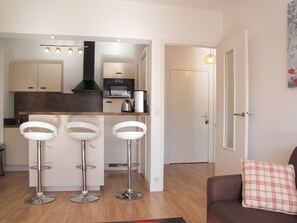 A modern kitchen with a built-in oven, a coffee machine, and a black range hood. There are three white bar stools and a brown sofa with a plaid pillow. - Homerez - Apartment in Antibes (Antibes)