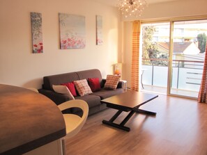 A modern living room with a sofa, a coffee table, and a dining area. There are paintings on the wall and a view of the outside through large windows. - Homerez - Apartment in Antibes (Antibes)