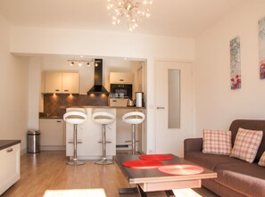 A modern kitchen with a central island, bar stools, a dining table with red placemats, and a living area with a sofa and decorative pillows. - Homerez - Apartment in Antibes (Antibes)