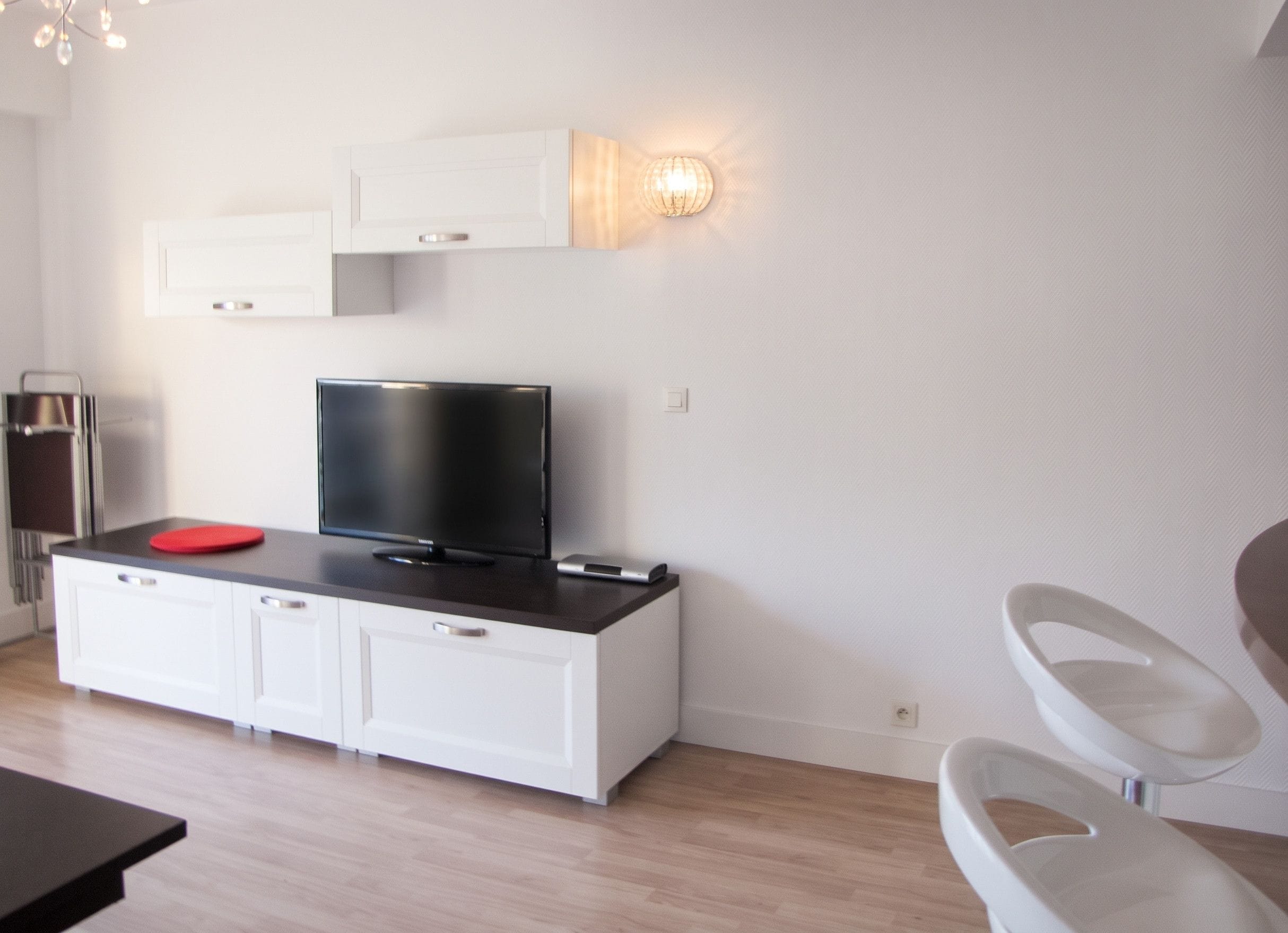 A modern living room with a flat-screen TV on a white cabinet, a red circular object on the cabinet, and a wall-mounted light fixture.