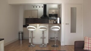 A modern kitchen with a central island, three bar stools, a trash can, and a door with a window. - Homerez - Apartment in Antibes (Antibes)