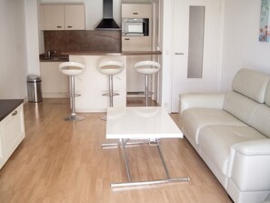 A modern kitchen with a built-in oven, a dining area with a white table and three bar stools, and a white sofa in a living area. - Homerez - Apartment in Antibes (Antibes)