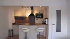 A compact kitchen with a central island, two bar stools, a stainless steel trash can, and a modern chandelier. - Homerez - Apartment in Antibes (Antibes)