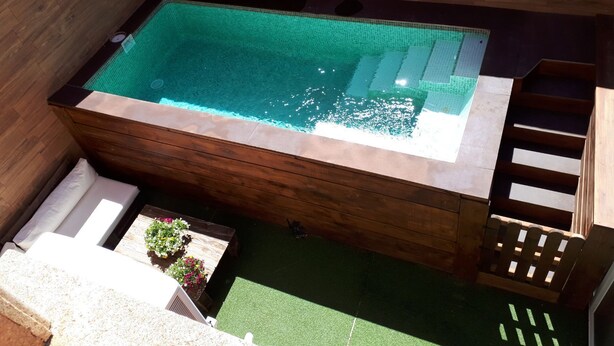 A rectangular hot tub with a wooden frame and a tiled interior, surrounded by a wooden deck and a small step leading to a wooden bench.