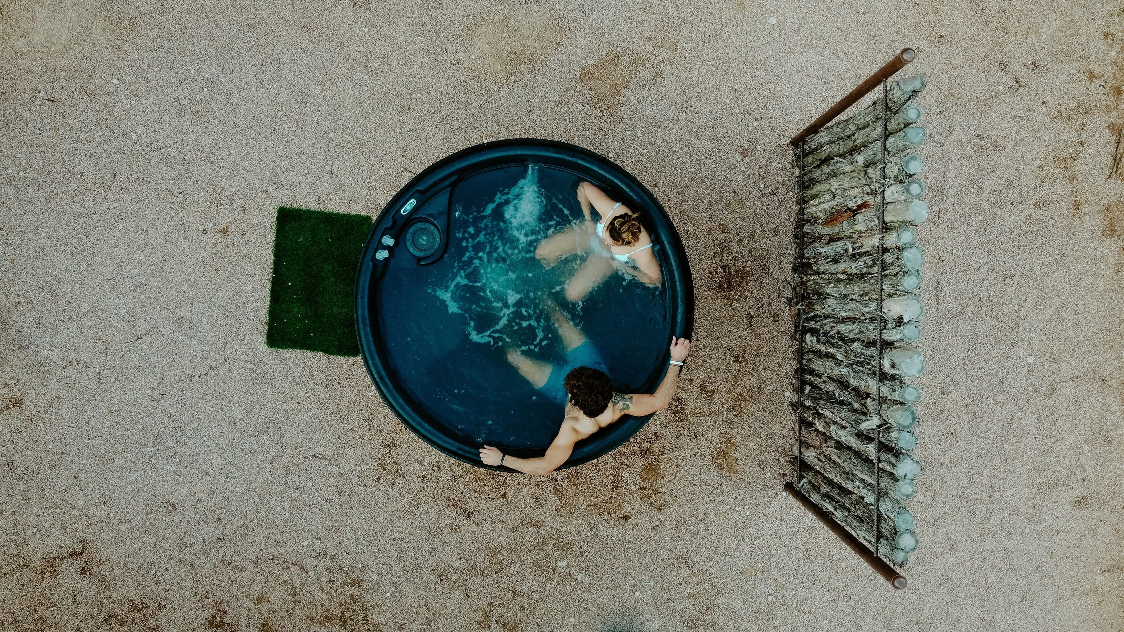 Una piscina circular con dos personas nadando, rodeada por una zona arenosa con un tapete verde y un estante metálico con botellas.