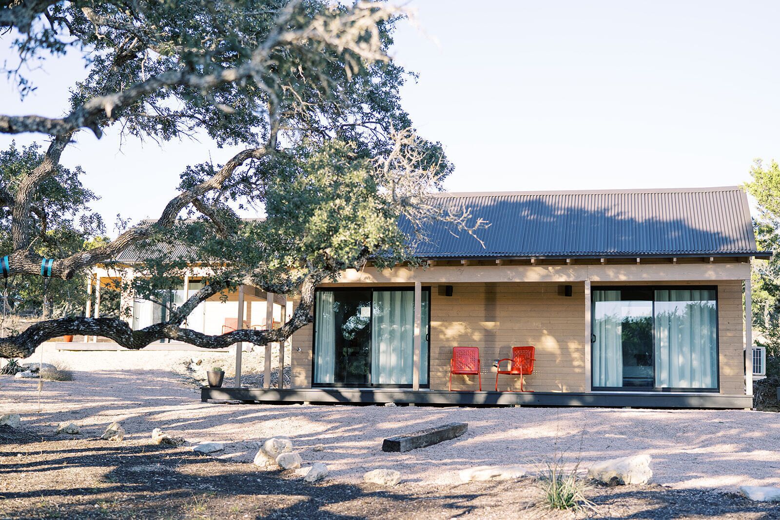 A modern house with a large window, a patio with two red chairs, and a tree in the foreground.