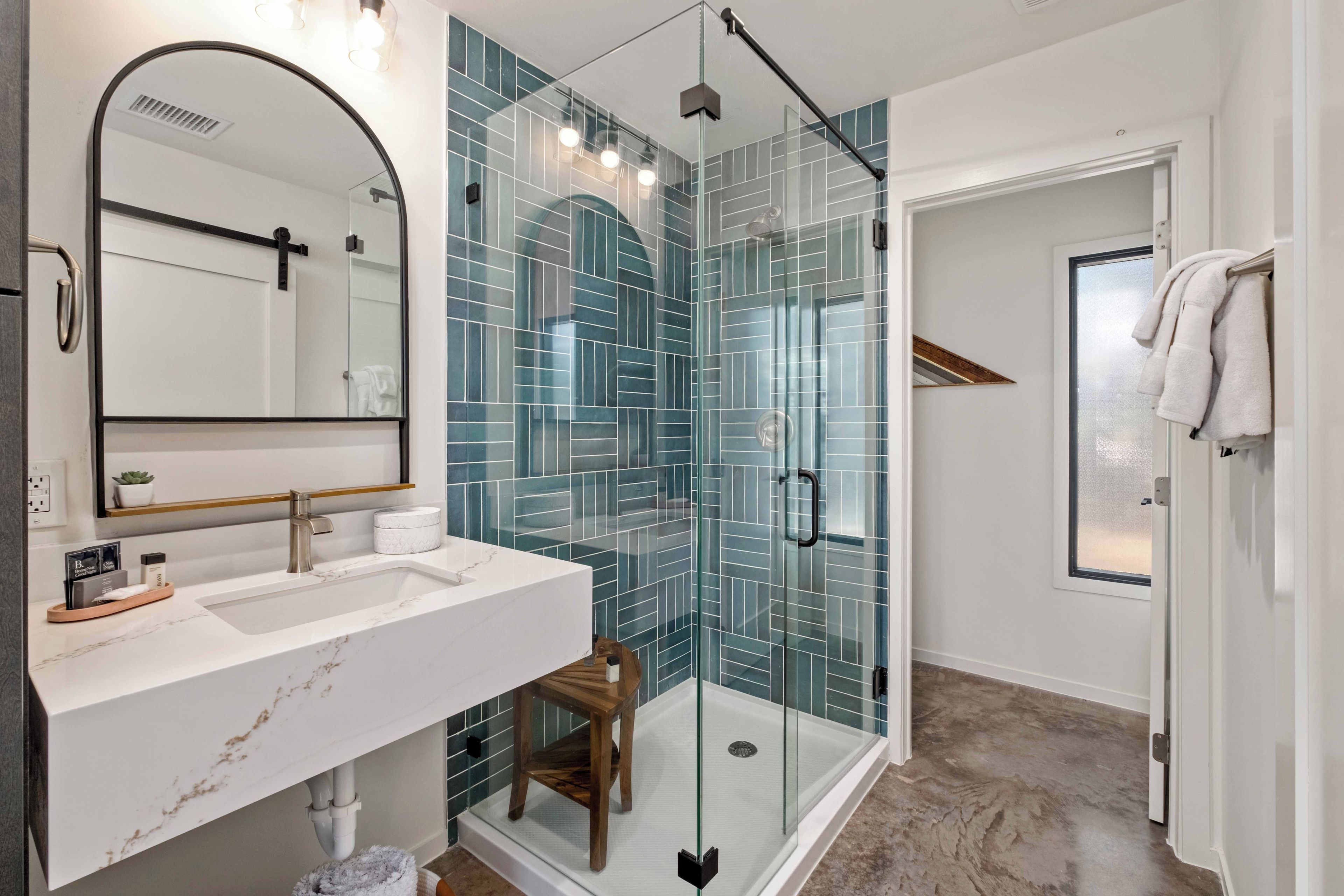 Un bany modern amb dutxa protegida per vidre, un lavabo blanc i una superfície de marbre, i un mirall gran.