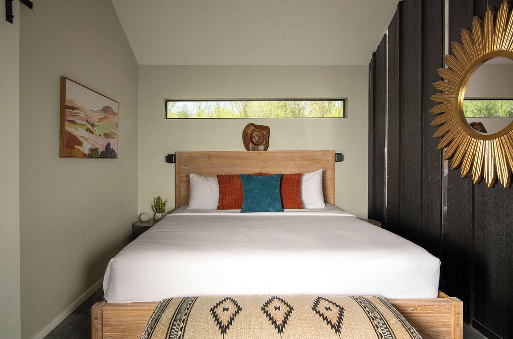 A bedroom with a large bed, a window, a wall-mounted mirror, and a painting.