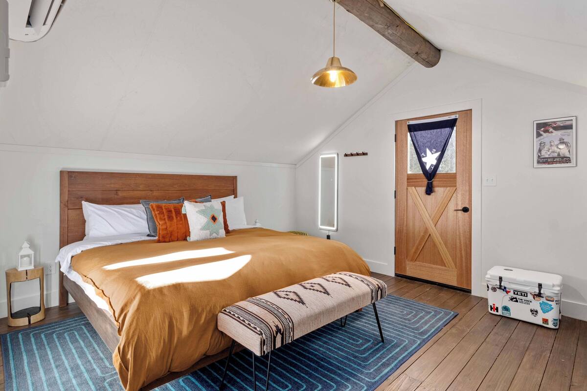 A bedroom with a large bed, a wooden headboard, a bench, a blue rug, a wooden door, and a storage box.