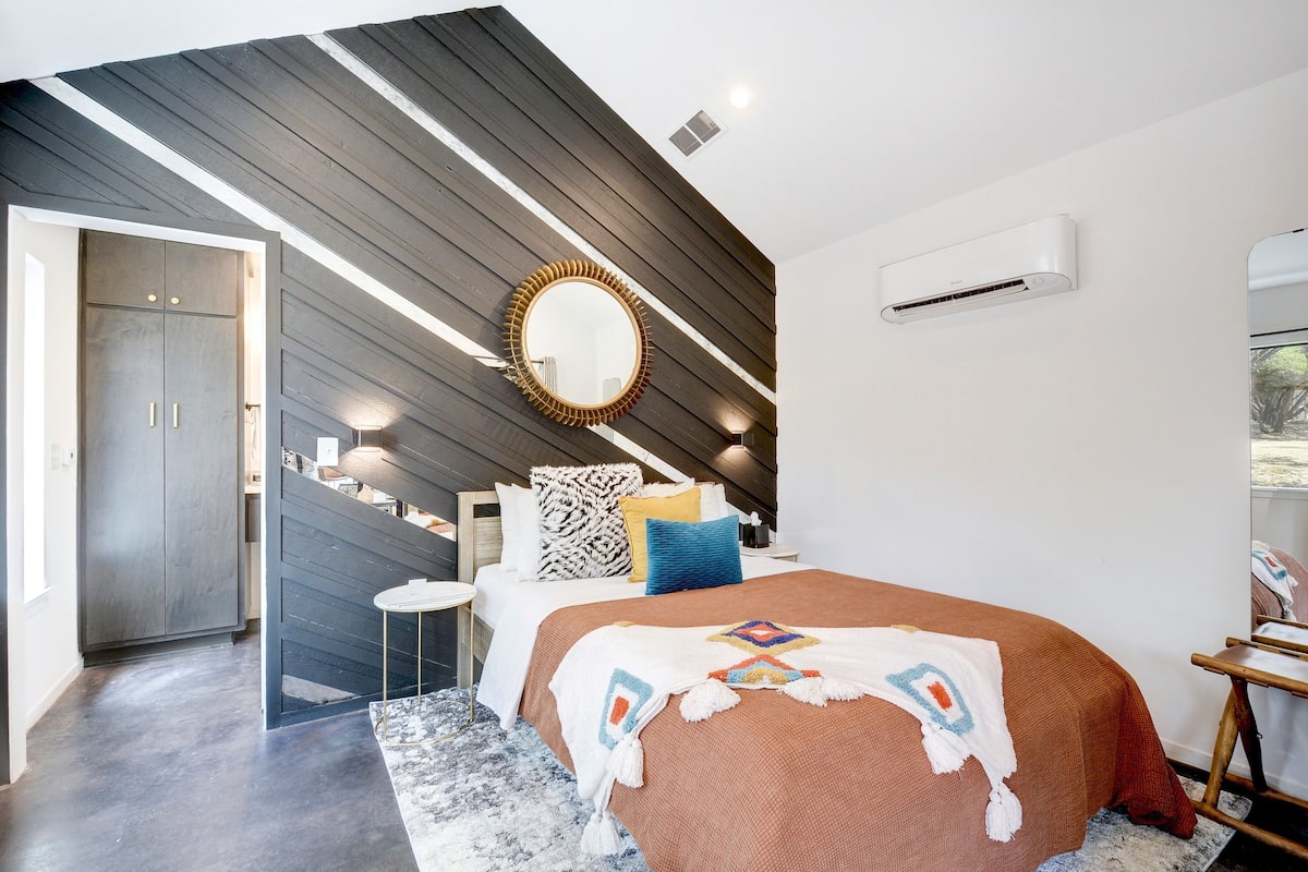 A modern bedroom with a sloped ceiling, a large bed with a patterned throw, a round mirror, and a small side table.