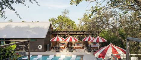 A pool area with lounge chairs and umbrellas, surrounded by trees and a building with a sloped roof.