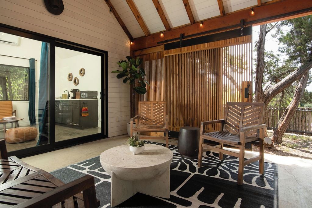A modern outdoor seating area with a round table, two wooden chairs, and a large window overlooking a kitchen.