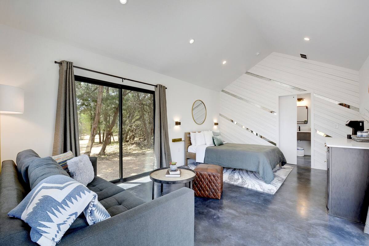 A modern, well-lit room with a sofa, a bed, a small table, and a view of the outdoors.