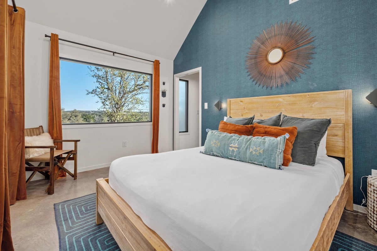 A bedroom with a large bed, a view of the outdoors, a wooden headboard, and a blue accent wall.