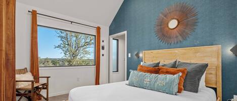 A bedroom with a large bed, a view of the outdoors, a wooden headboard, and a blue accent wall.