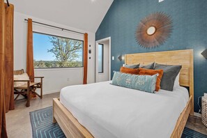 A bedroom with a large bed, a view of the outdoors, a wooden headboard, and a blue accent wall. - Lucky Arrow Retreat - Dripping Springs (Dripping Springs)