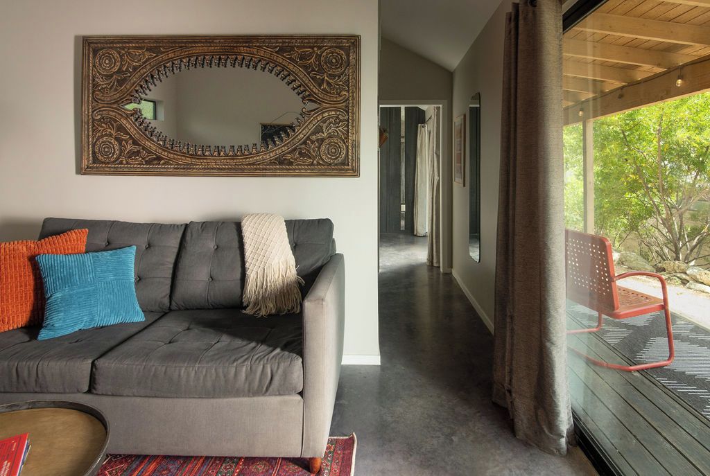 Un salon moderne avec un canapé gris, un grand miroir et une vue sur un jardin, visible à travers une porte vitrée.