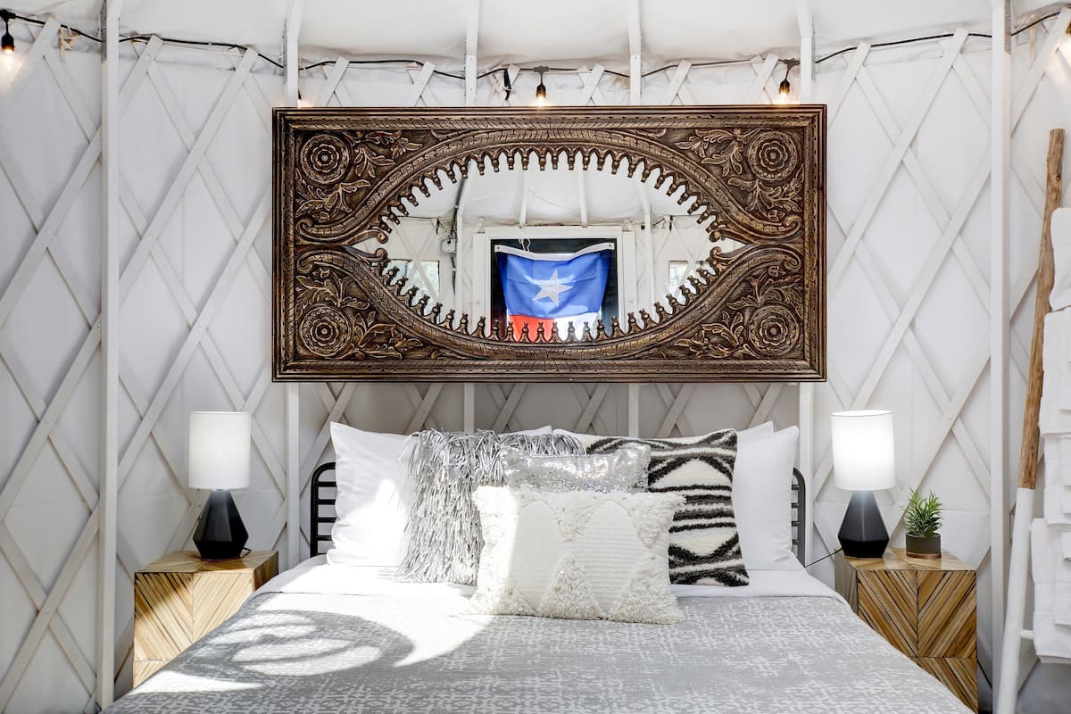 A bedroom with a large ornate mirror, a bed with patterned bedding, two bedside tables with lamps, and a small plant on one table.