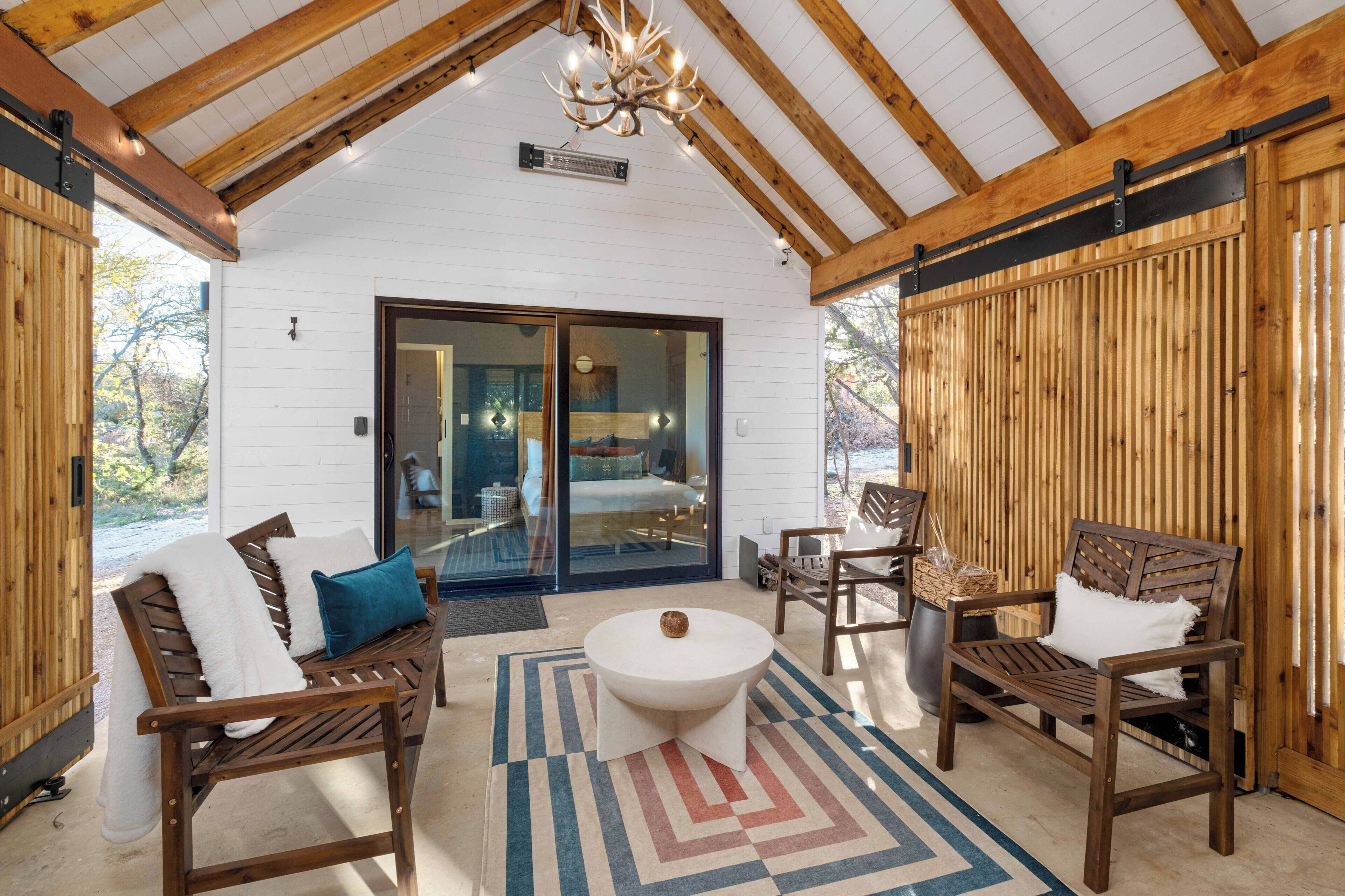 A cozy outdoor seating area with wooden furniture, a round table, and a view of a bedroom through glass doors.
