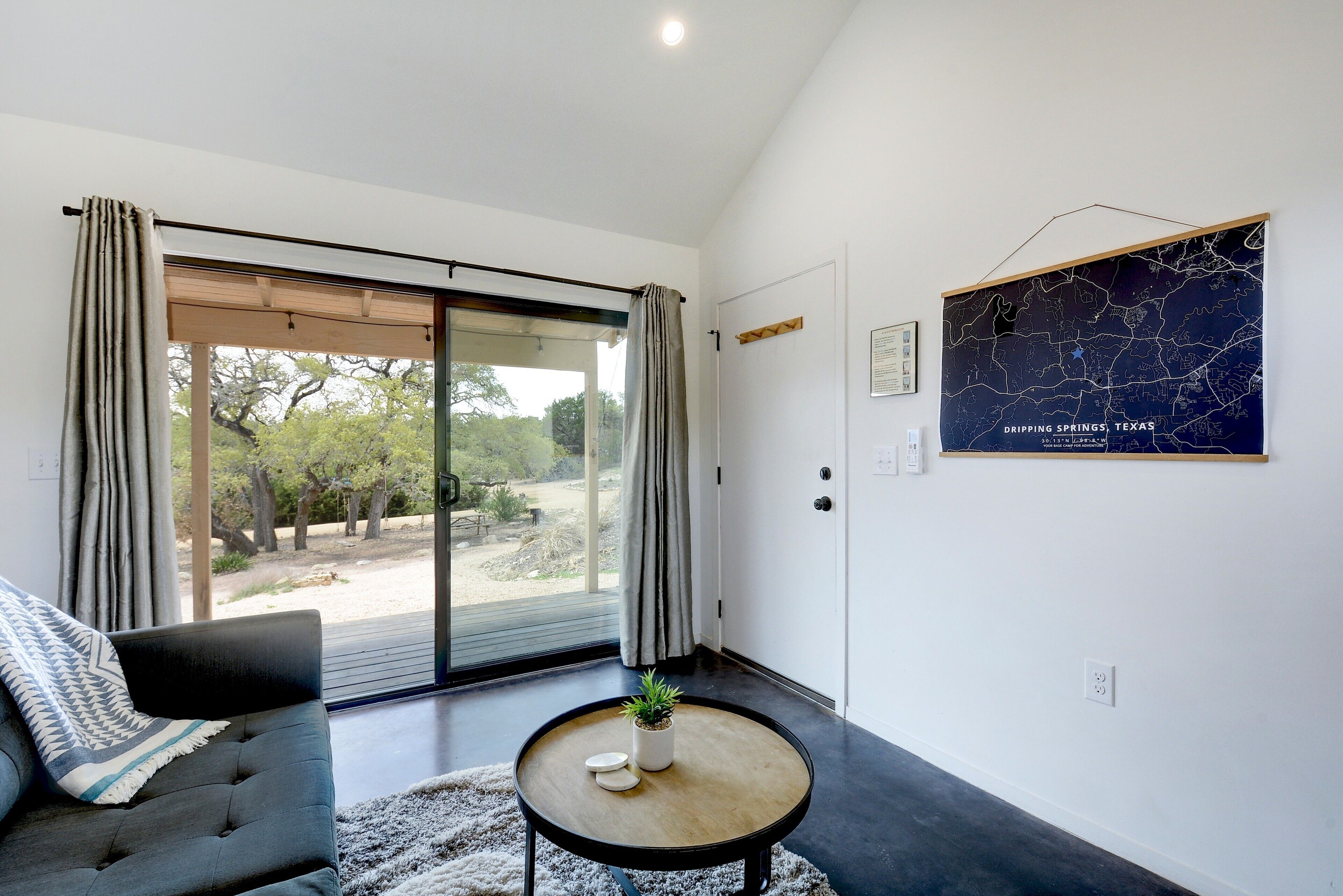 A modern living room with a large sliding glass door leading to an outdoor area, a dark sofa with a throw blanket, a small round table with a plant, and a map of Texas on the wall.