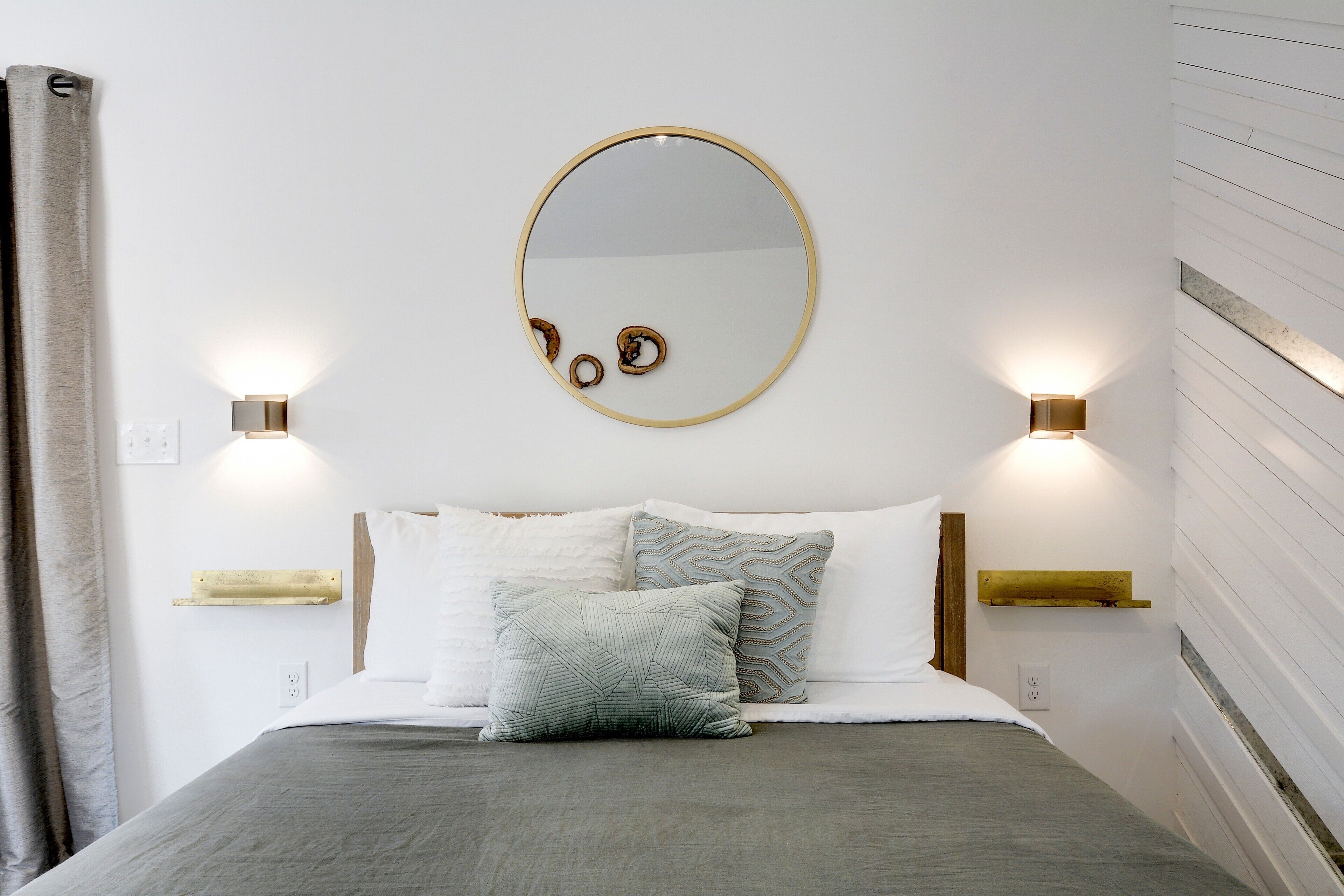 A bedroom with a bed, pillows, a round mirror, and wall-mounted lights.