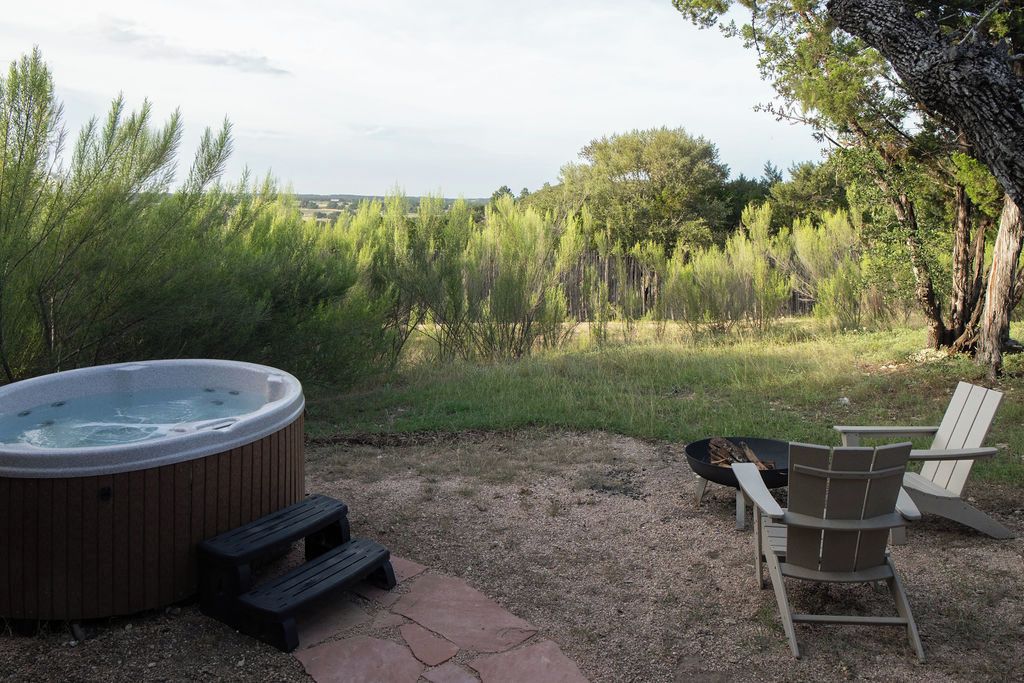 A hot tub, two Adirondack chairs, and a fire pit in a backyard with trees and a grassy field.