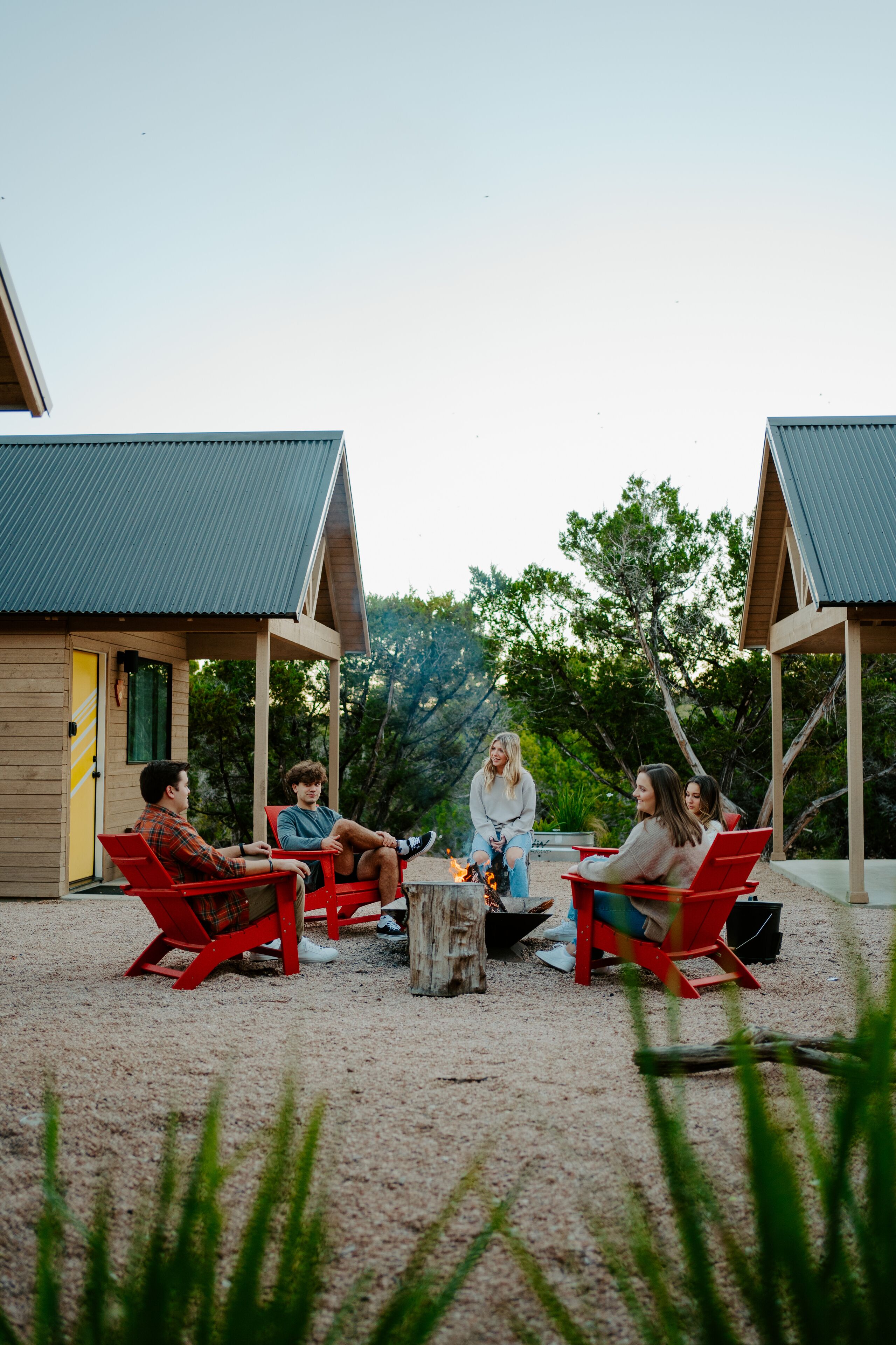 A group of people sitting around a fire pit in a covered outdoor area with wooden cabins in the background.