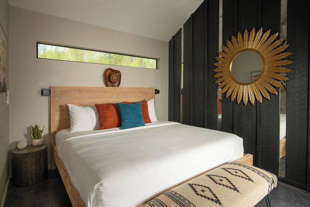 A modern bedroom with a wooden headboard, a large mirror, a window, and a bedside plant.