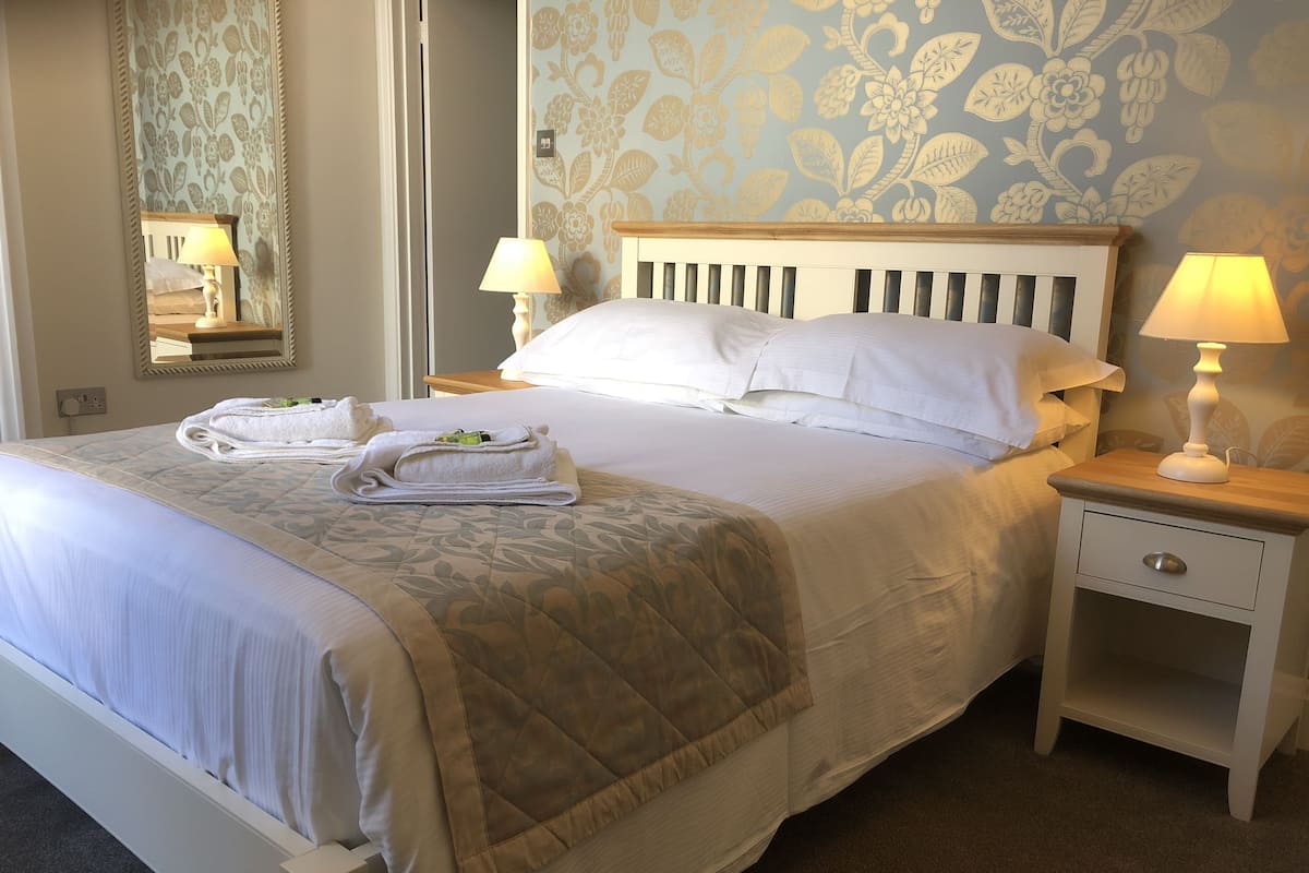 A neatly made bed with a quilted cover, two bedside lamps, a wooden nightstand with a drawer, and a mirror on the wall.