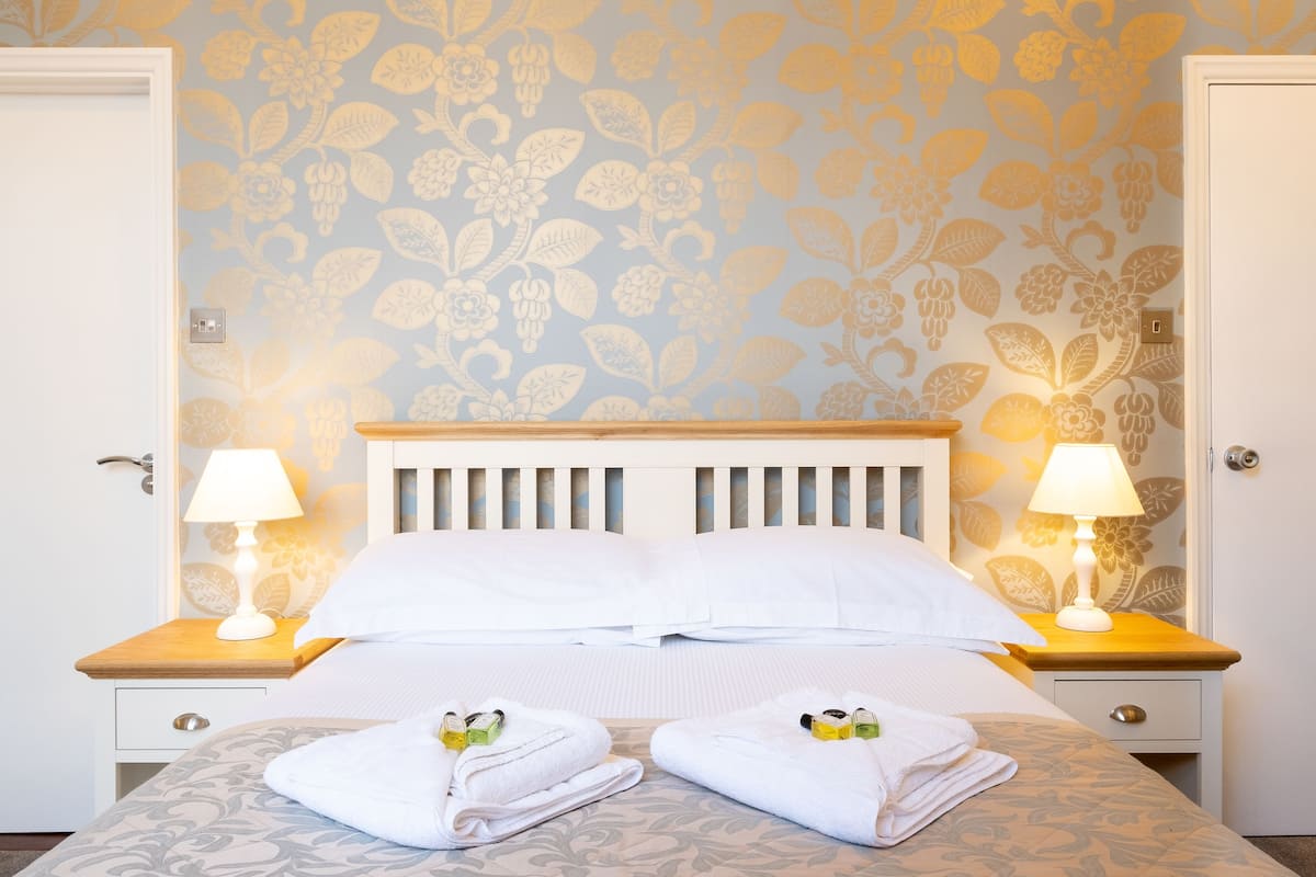 A neatly made bed with white linens, two bedside lamps, and a floral wallpaper pattern.
