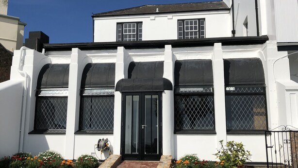 Un edificio blanco con una puerta negra, varias ventanas con rejas de metal y un camino de piedra que conduce a la entrada.