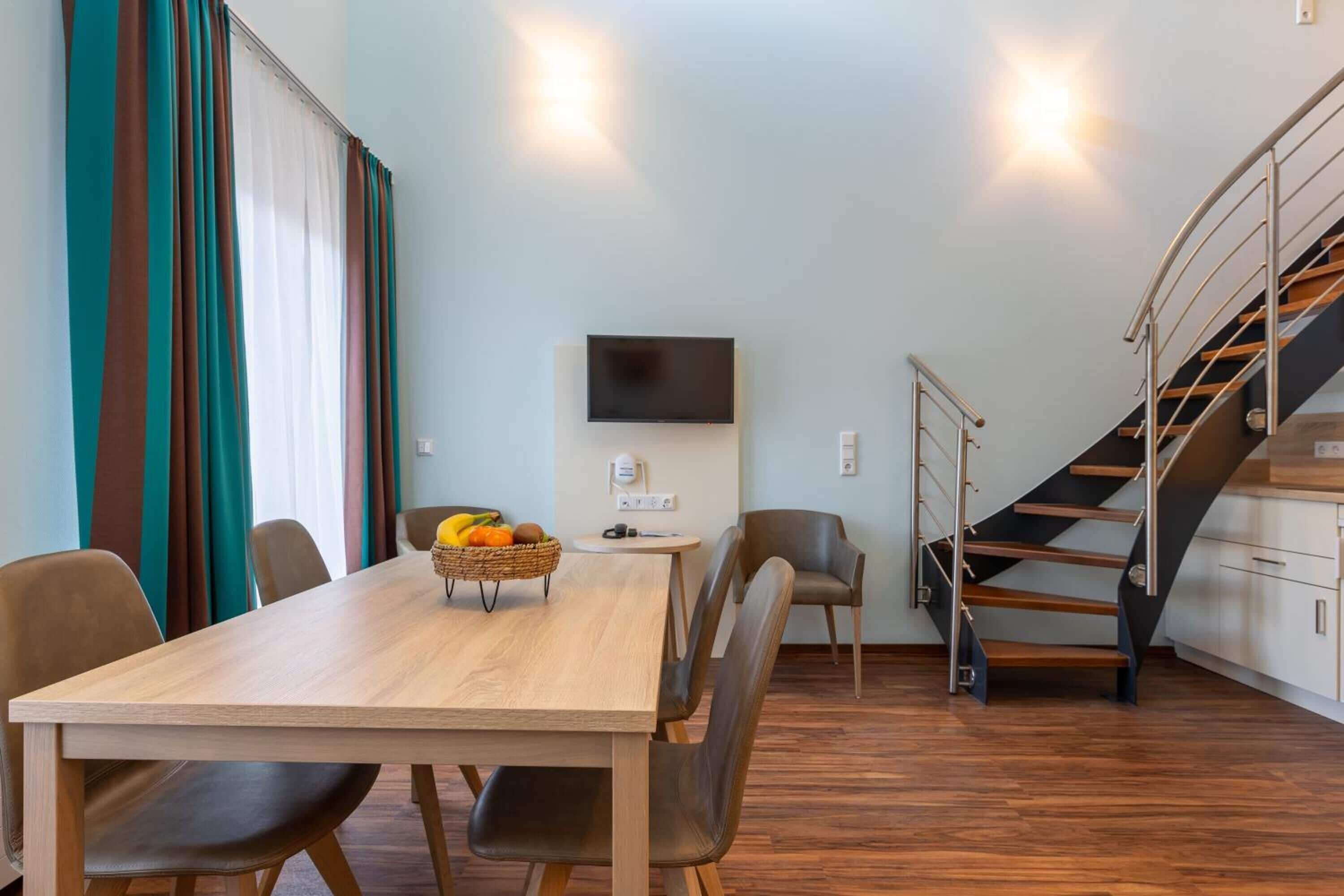 A modern living room with a wooden dining table, leather chairs, a TV mounted on the wall, and a staircase with a metal railing.