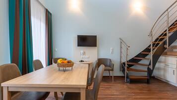 A modern living room with a wooden dining table, leather chairs, a TV mounted on the wall, and a staircase with a metal railing.