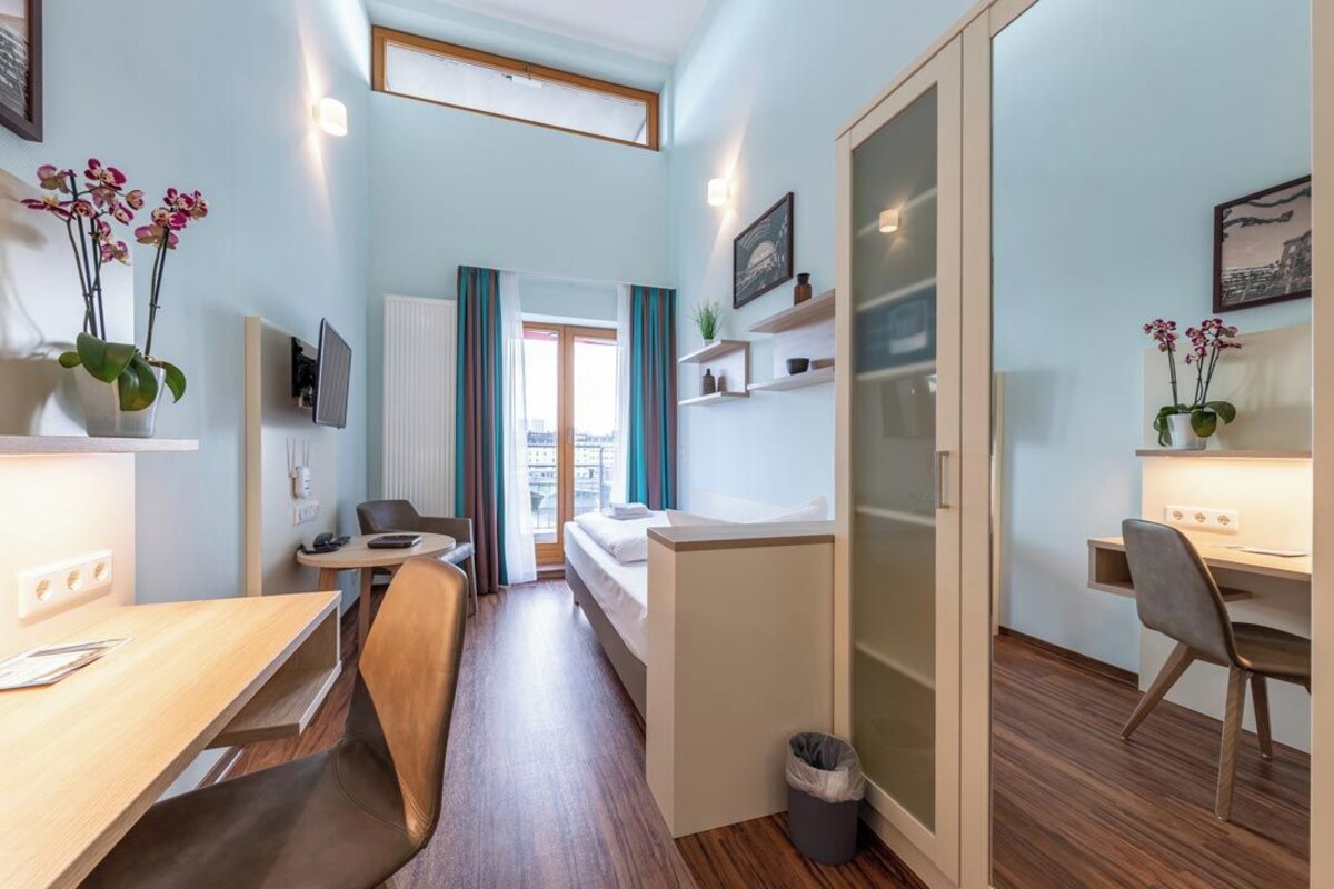 A compact hotel room with a bed, a desk, a chair, a small table, and a trash bin. There are potted plants and a TV on the wall.