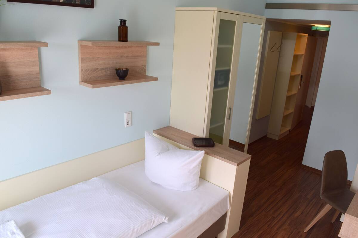 A small, neatly arranged bedroom with a single bed, a bedside table, a wardrobe, and a chair.