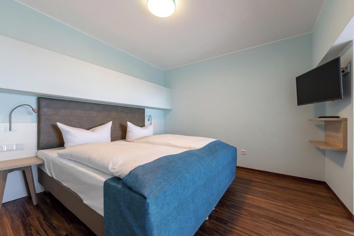 A modern hotel room with a large bed, a bedside table, a flat-screen TV mounted on the wall, and a wooden floor.