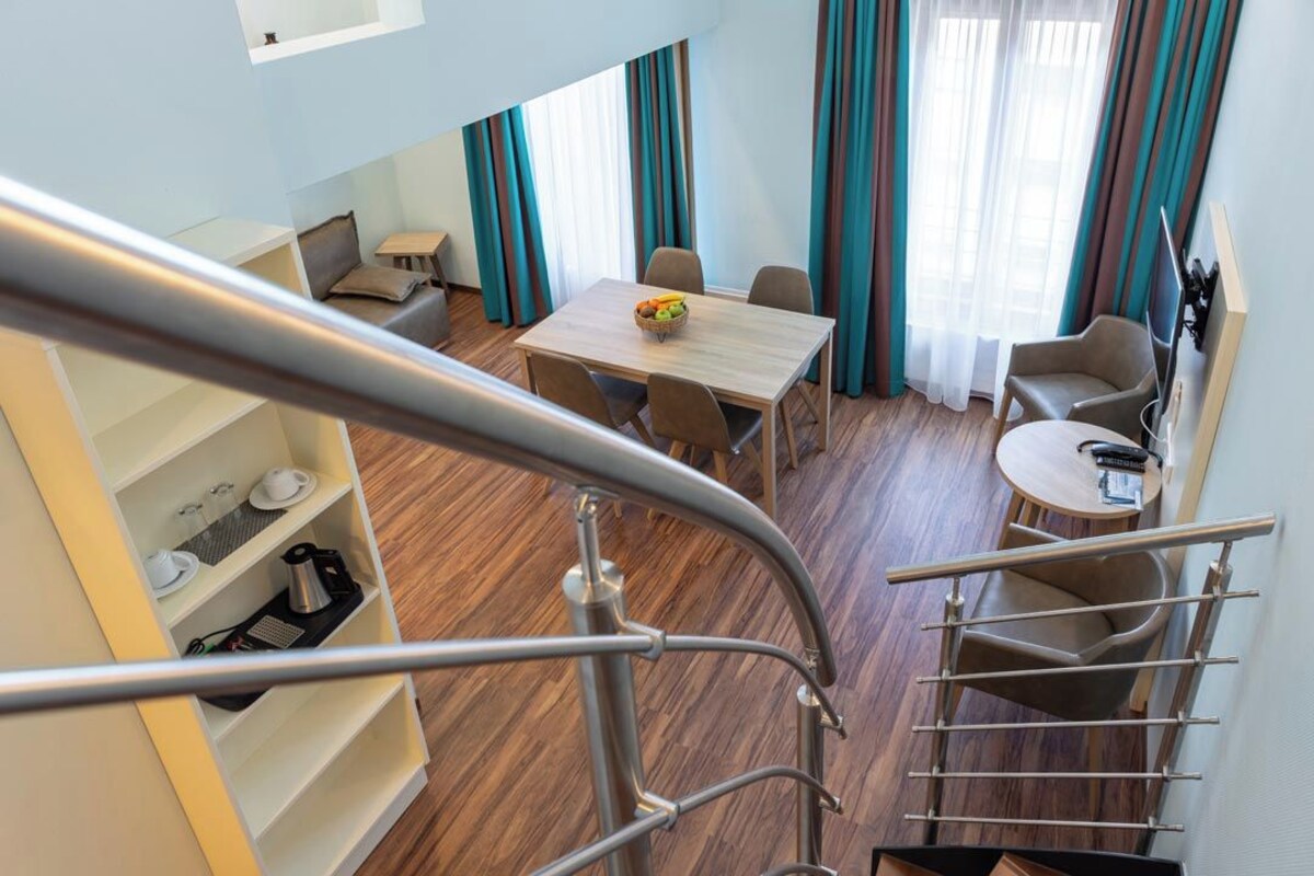 A modern hotel room with a dining area, a small kitchen, and a staircase.