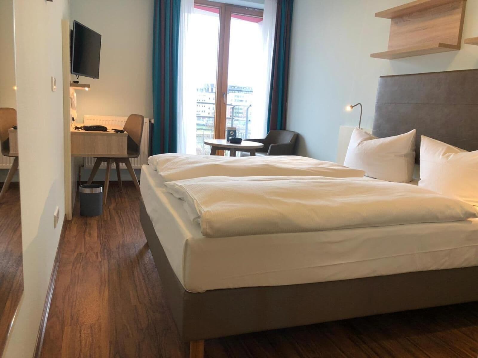 A hotel room with a large bed, a desk with a chair, a TV mounted on the wall, and a view of the city through the window.