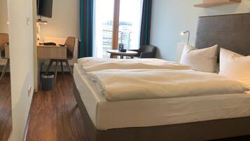 A hotel room with a large bed, a desk with a chair, a TV mounted on the wall, and a view of the city through the window.